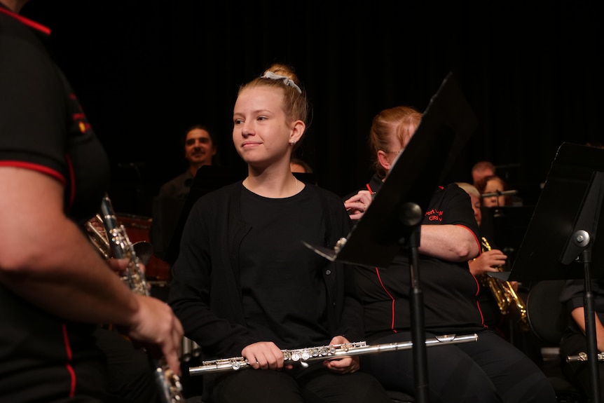 A young firl with a flute in her lap smiles at other orchestra members.