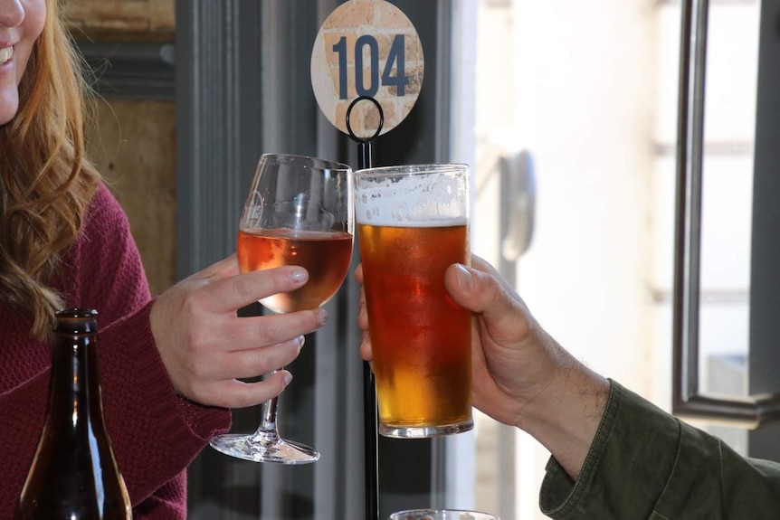 A couple gently clink their wine and beer glasses together at a pub table.