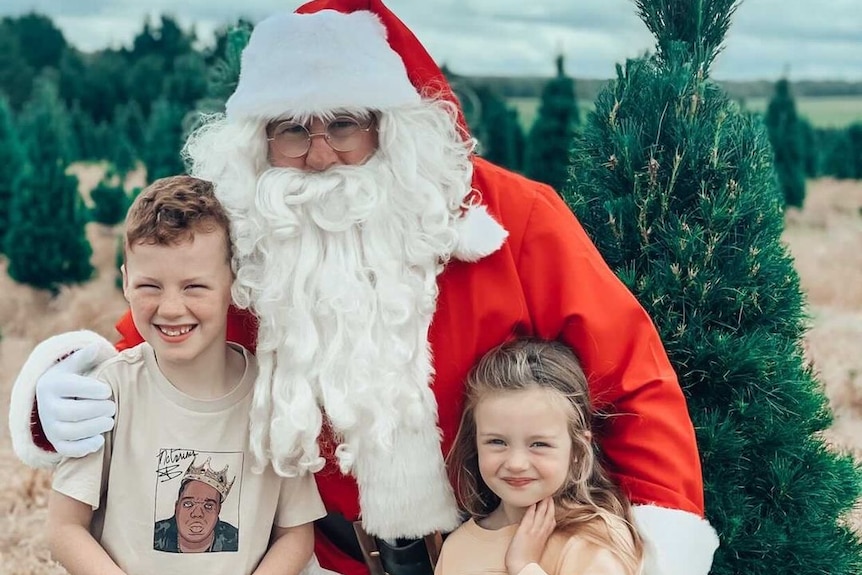 Two children are being hugged by Santa on a Christmas tree farm