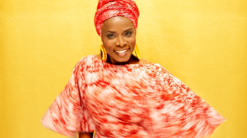Angélique Kidjo spins against a yellow background wearing red and white traditional African garments