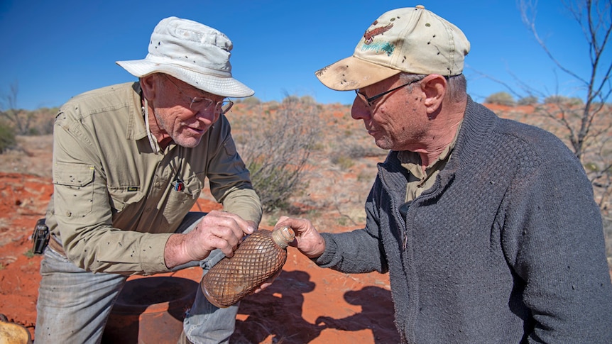 Two men crouch down in the Simpson desert and examine old bottle with flies around them