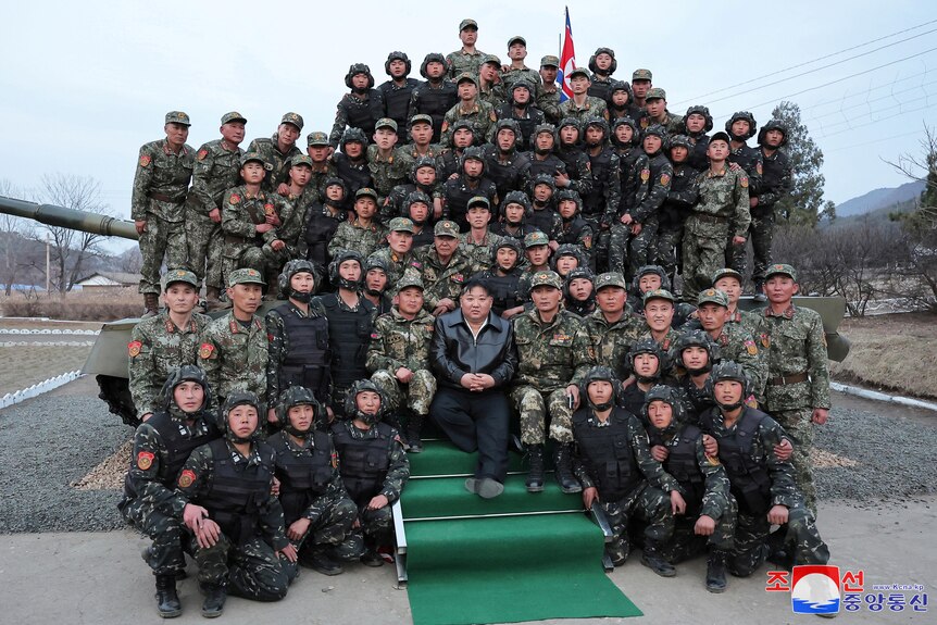 North Korean leader Kim Jong Un poses with uniformed soldiers, sitting in rows in front of a tank. 