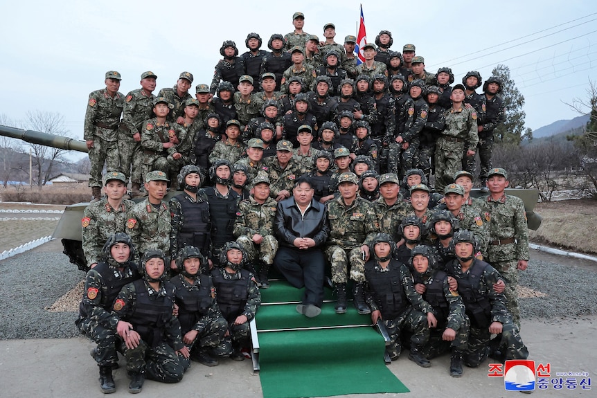 North Korean leader Kim Jong Un poses with uniformed soldiers, sitting in rows in front of a tank. 