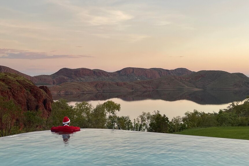 a man in a santa suit leans on the edge of Lake Argyle's iconic infinity pool looking at ref cliffs in the distance