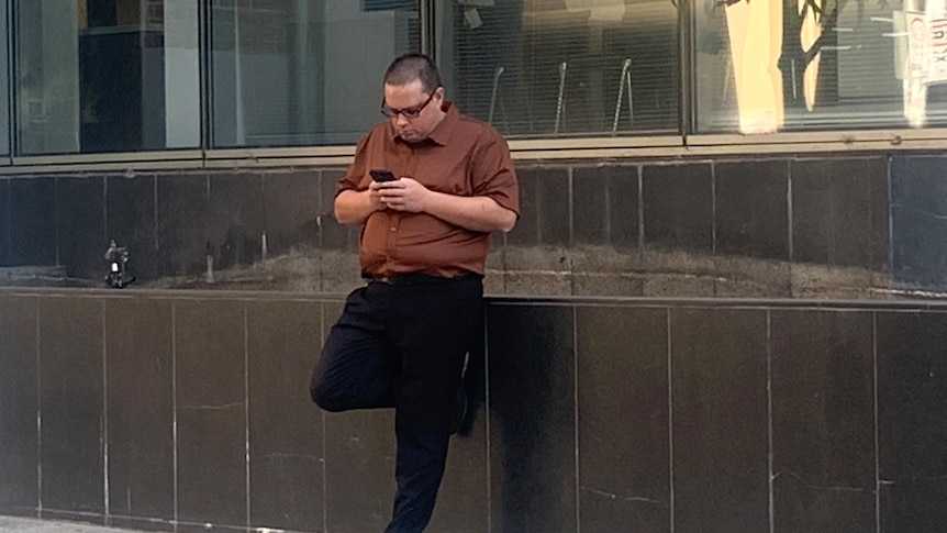 A man named Travis Barnard stands leaning against a wall in black pants and a brown shirt, typing on his mobile phone.