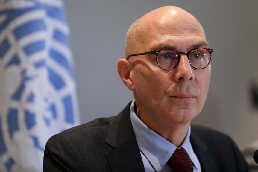 Bald man with glasses sitting with UN flag behind him