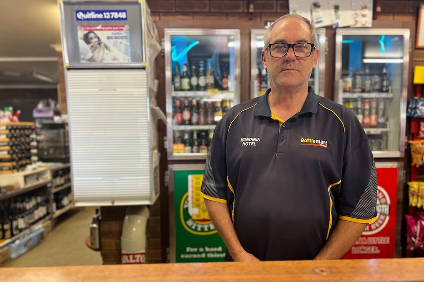 A man wearing glasses stands inside a bottle shop. 