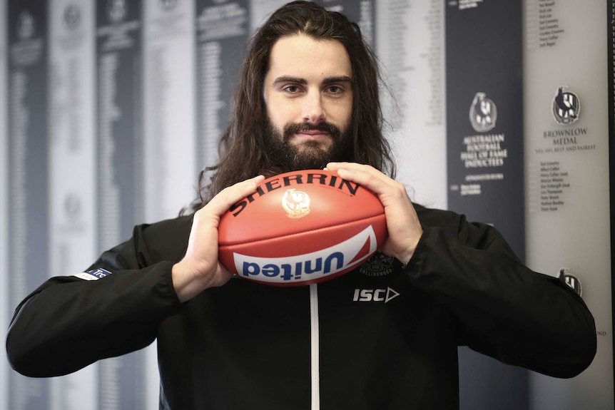 Brodie Grundy holds a football while posing for a photo. He's in a Collingwood jacket in front of a club honours board.