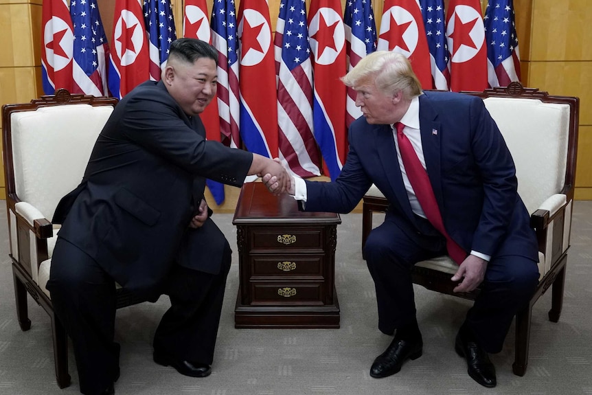 Two men shaking hands while seated before a row of North Korean and US flags