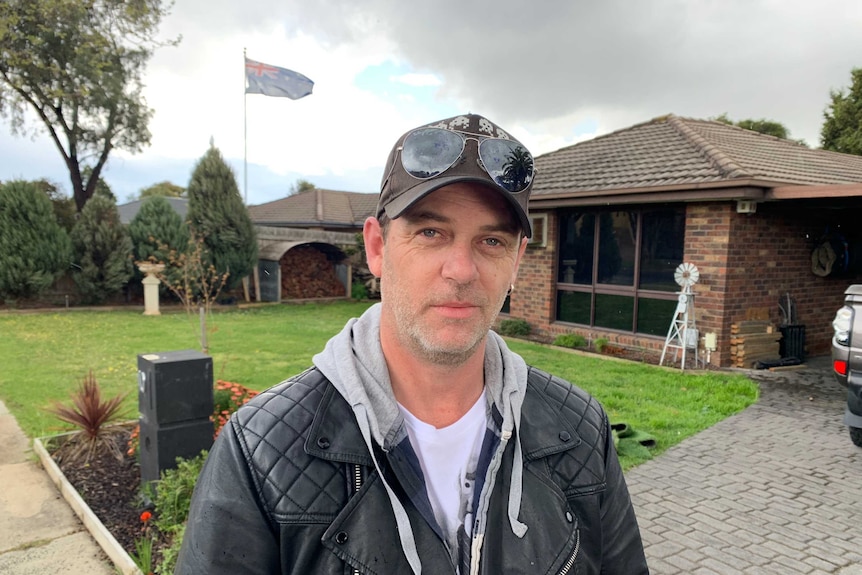 Dave Chapman, in a black jacket, cap and sunglasses on his head, in front of a brick home.