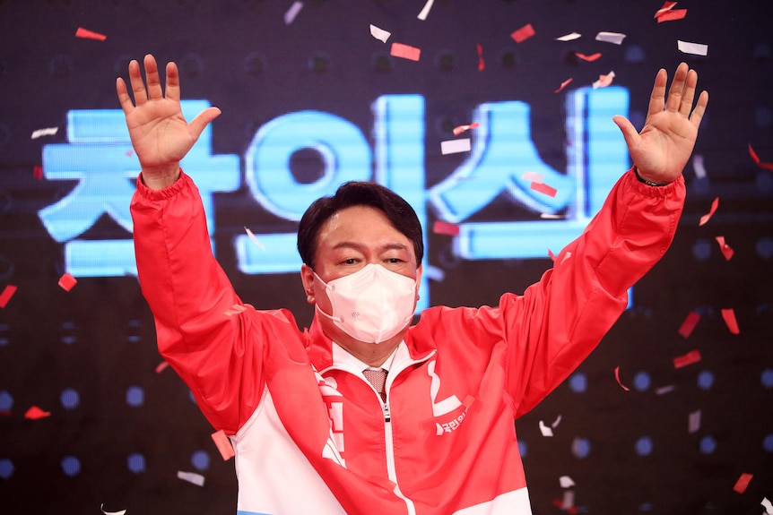 Yoon Suk-yeol wearing a red and white windcheater and white mask raises his hands in the air as confetti rains down on sta