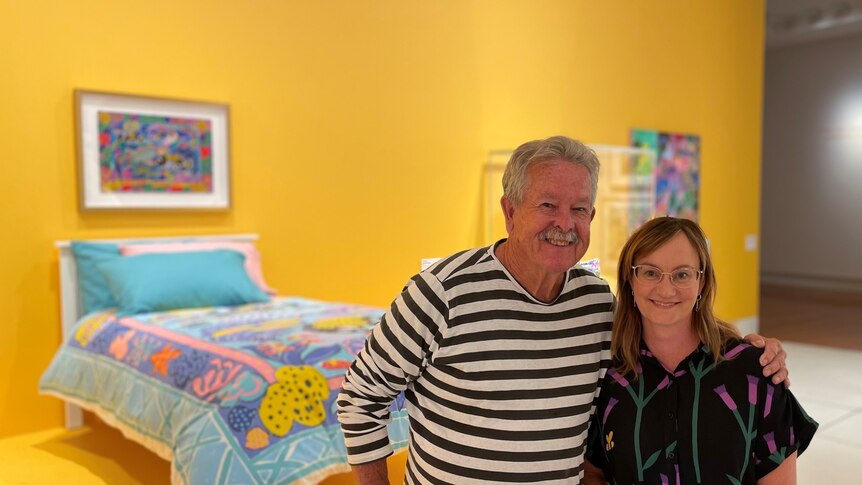 a man and woman stand in front of a colourful bed set in a gallery