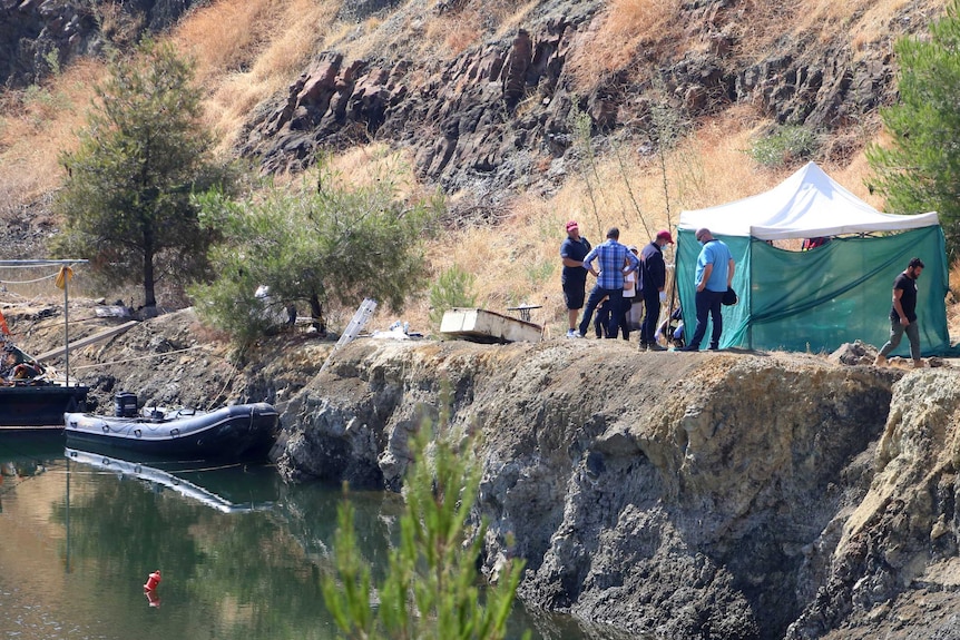 A small boat is docked by a lake with rocks and shrubs around as men stand nearby a makeshift tent.