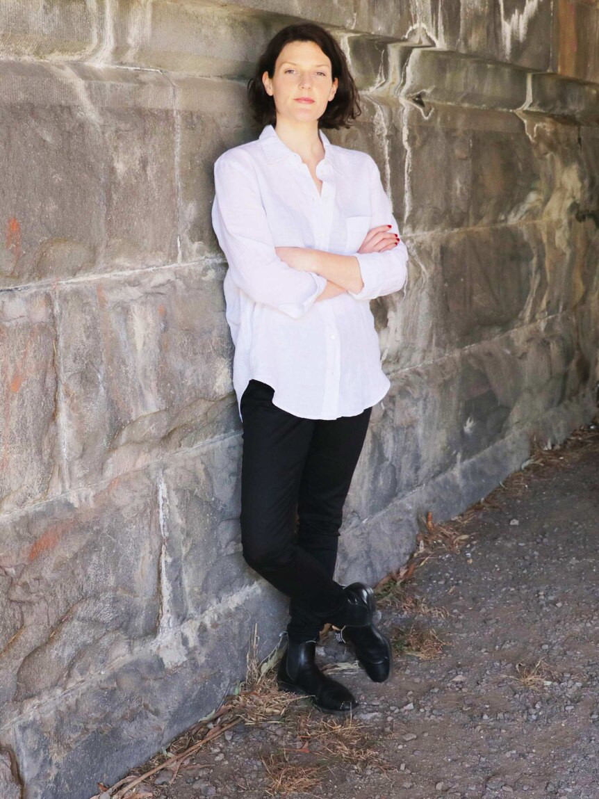 Sigrid McCarthy stands against a wall, wearing black jeans and a white shirt to depict a capsule wardrobe.