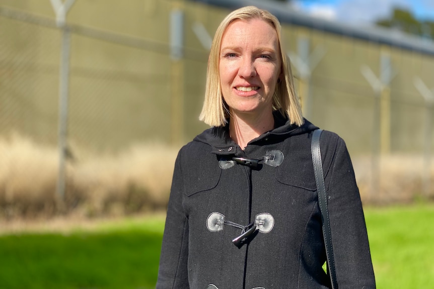 A blonde-haired woman with a black coat standing in front of a metal fence.