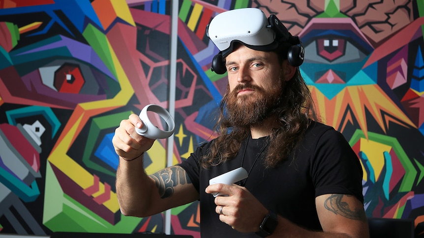 Ayjay holds two virtual reality controllers while wearing virtual reality goggles on top of his head, looking at the camera.