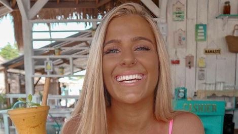 A tanned Chloe Roberts smiles as she holds a tropical drink in a bikini and short shorts.
