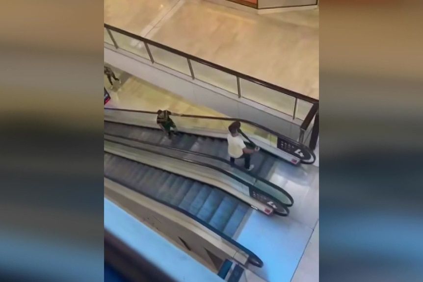 A man at the top of an escalator holds a bollard, to holds back a man with a knife