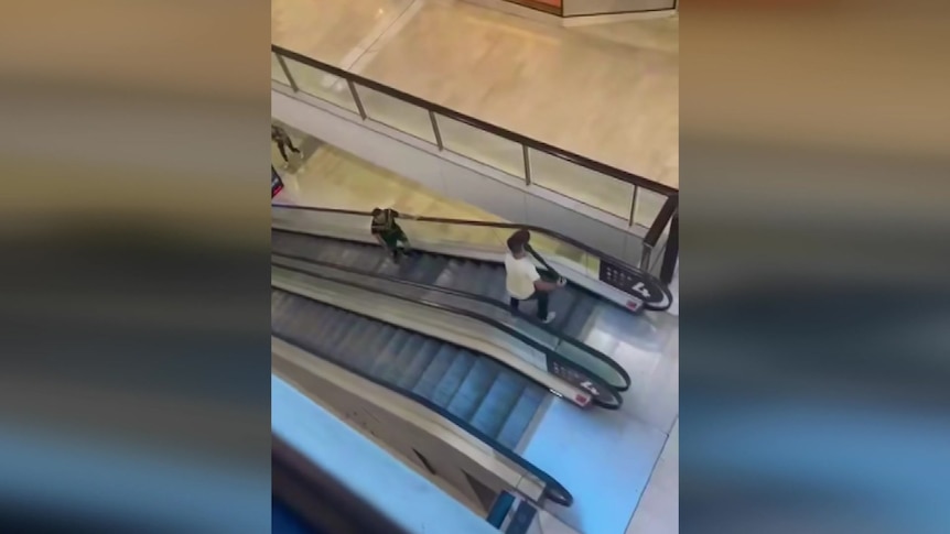 A man at the top of an escalator holds a bollard, to holds back a man with a knife