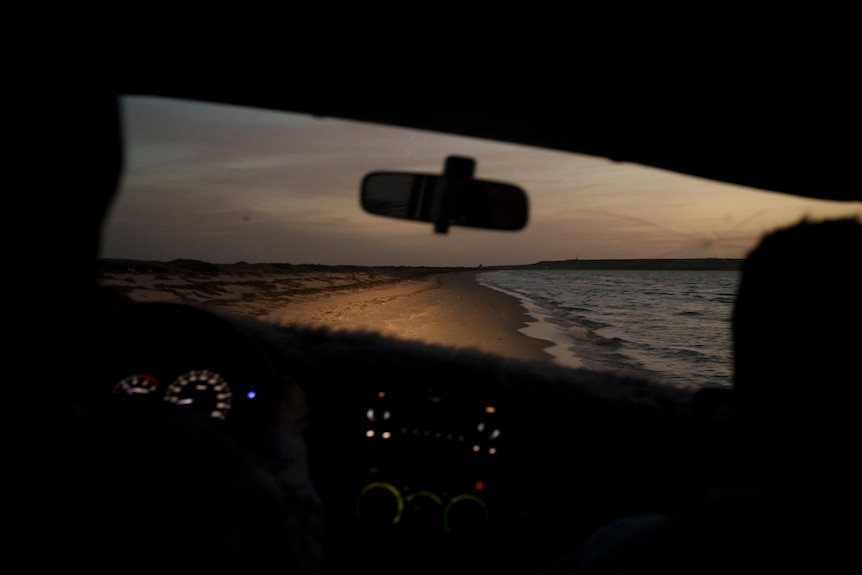 A beach is illuminated by a car's headlights at dusk.