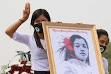 Two women in white hold three-finger salutes and an illustrated portrait of a teen killed in the protests