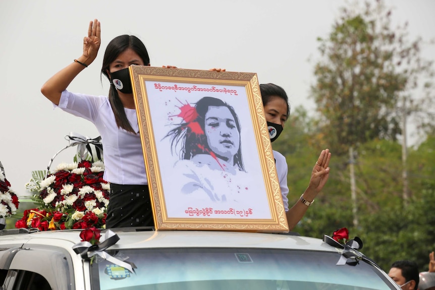 Two women in white hold three-finger salutes and an illustrated portrait of a teen killed in the protests