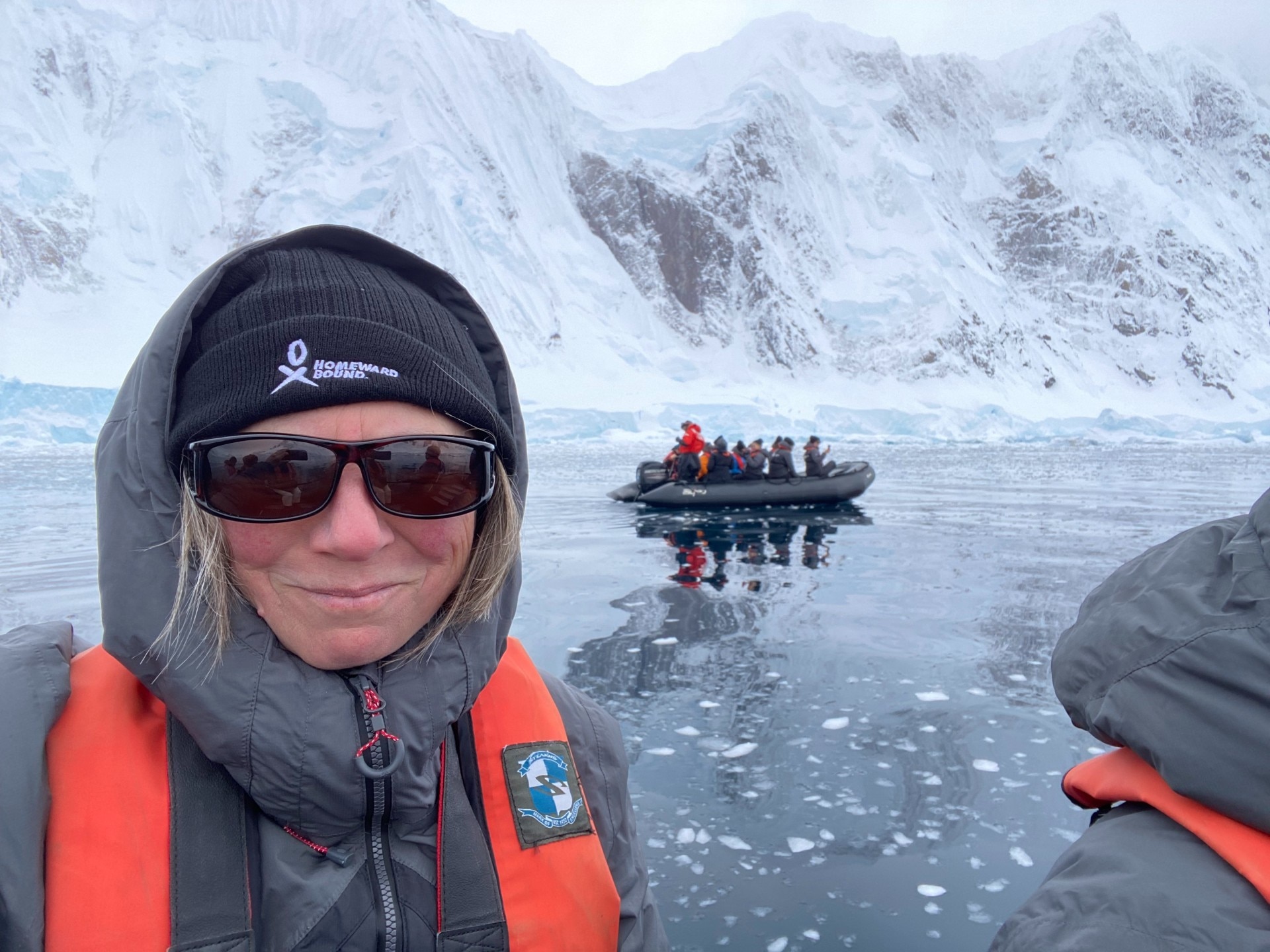 a woman in a black beanie sits in a dinghy. a grey hood pulled over her head, large black sunglasses and an orange life jacket