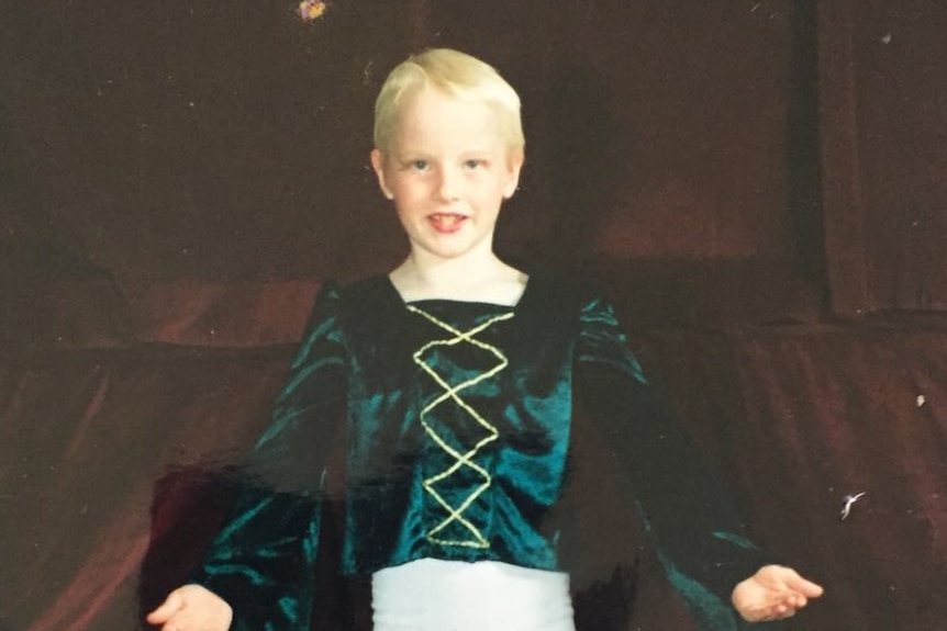 A young blonde boy in a dancing outfit in front of a curtain.