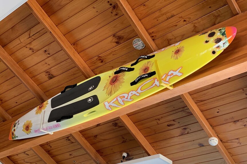 A yellow surfboard hangs near a wooden ceiling.