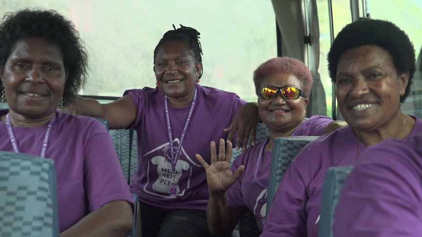 Four Papua New Guinean women in purple shirts sit in the back of a bus, smiling at the camera.