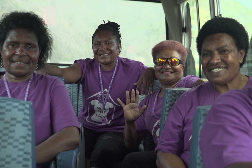 Four Papua New Guinean women in purple shirts sit in the back of a bus, smiling at the camera.