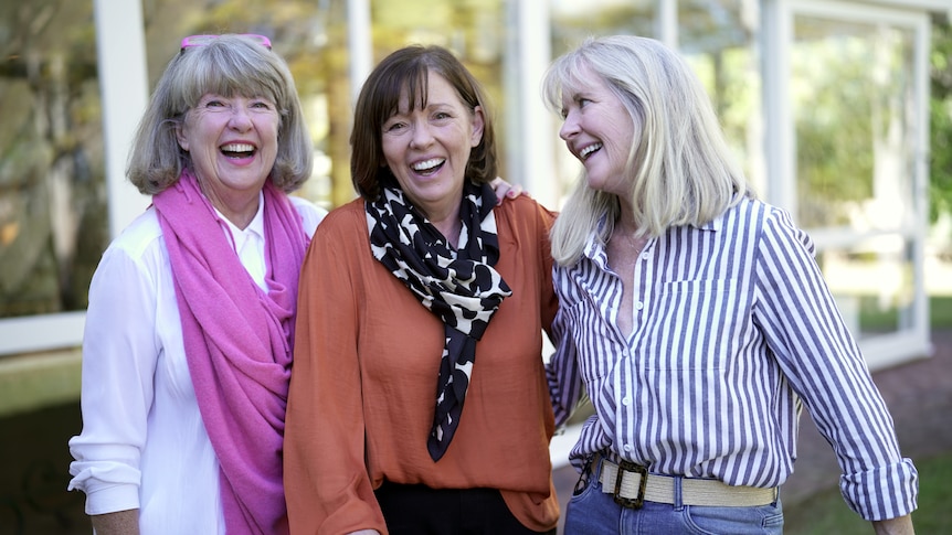 Three mature-aged women stand laughing and smiling