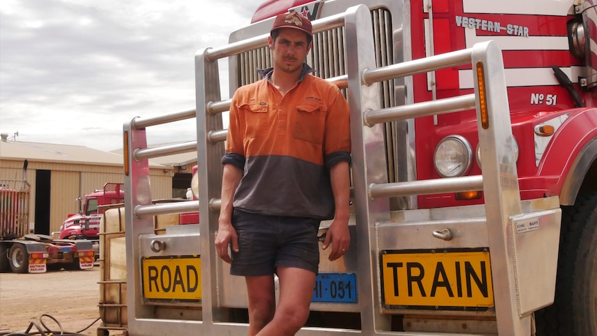 A young man with light facial hair, wearing high-vis, standing in front of truck.