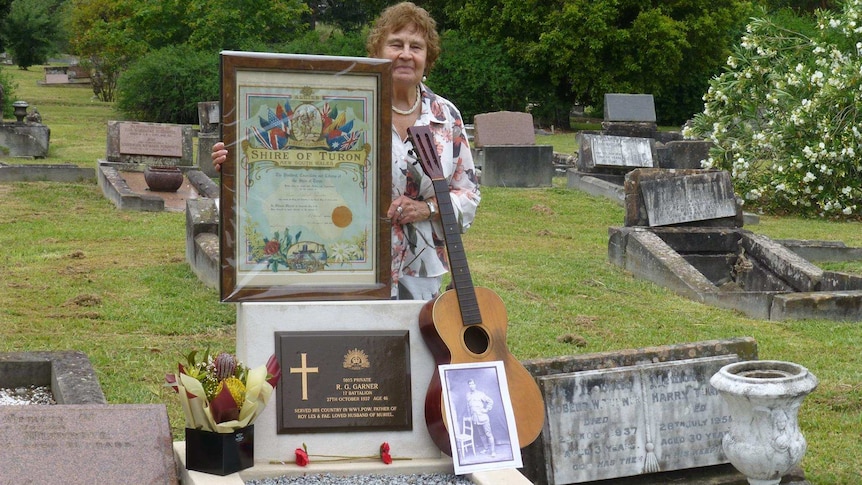 A woman stands at a grave, holding a framed certificate and old guitar.
