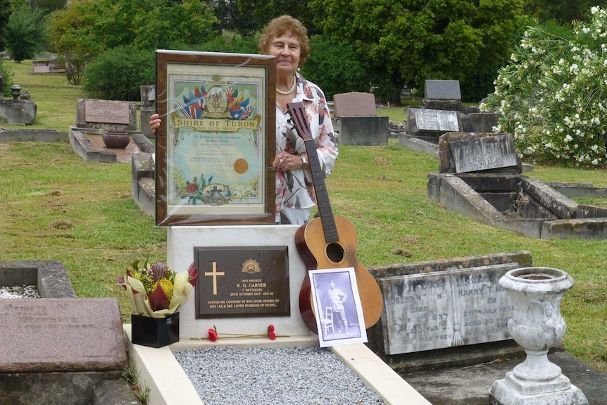 A woman stands at a grave, holding a framed certificate and old guitar.