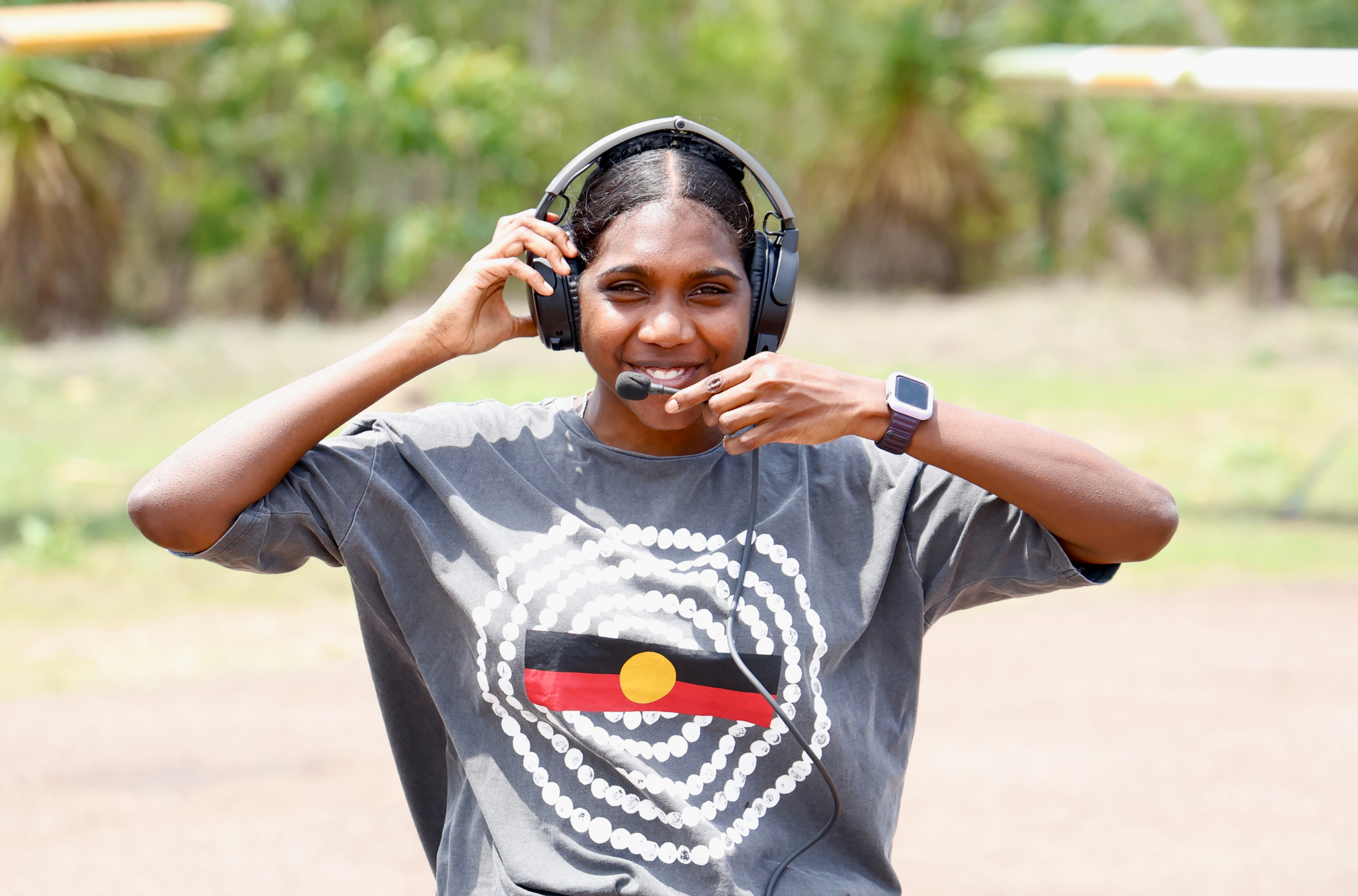 Smiling woman wearing t-shirt with Aboriginal flag stands on airstrip with her pilot's headphones