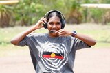 Smiling woman wearing t-shirt with Aboriginal flag stands on airstrip with her pilot's headphones