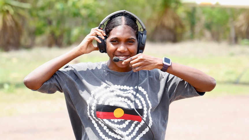 Smiling woman wearing t-shirt with Aboriginal flag stands on airstrip with her pilot's headphones