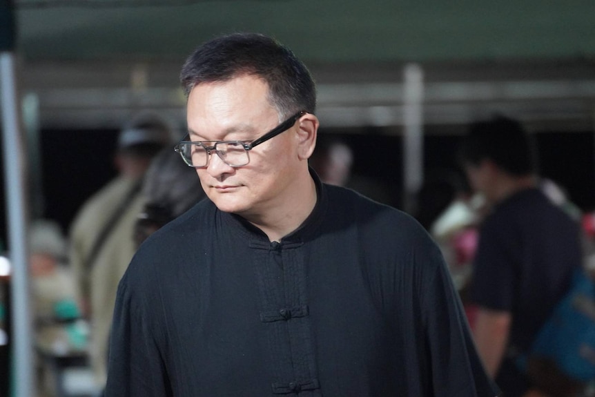 A middle aged bespectacled north Asian man wears a black shirt and looks away from the camera as he moves through a crowd.