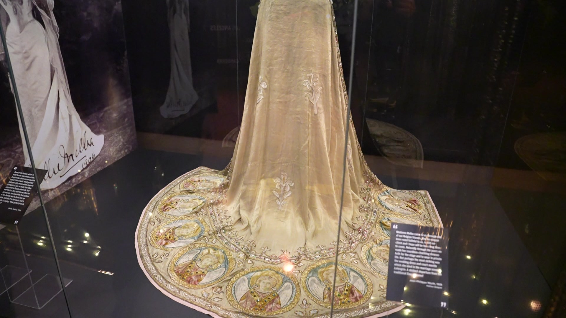 The bottom of a golden coloured cloak that is behind glass is spread out on the ground showing images of a woman.