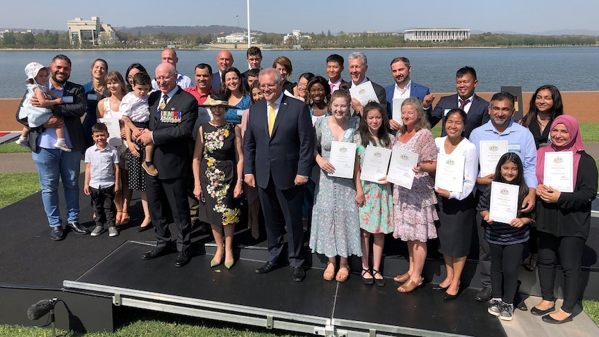 A group of people who have just attended their citizenship ceremony stand with Prime Minister Scott Morrison next to a lake