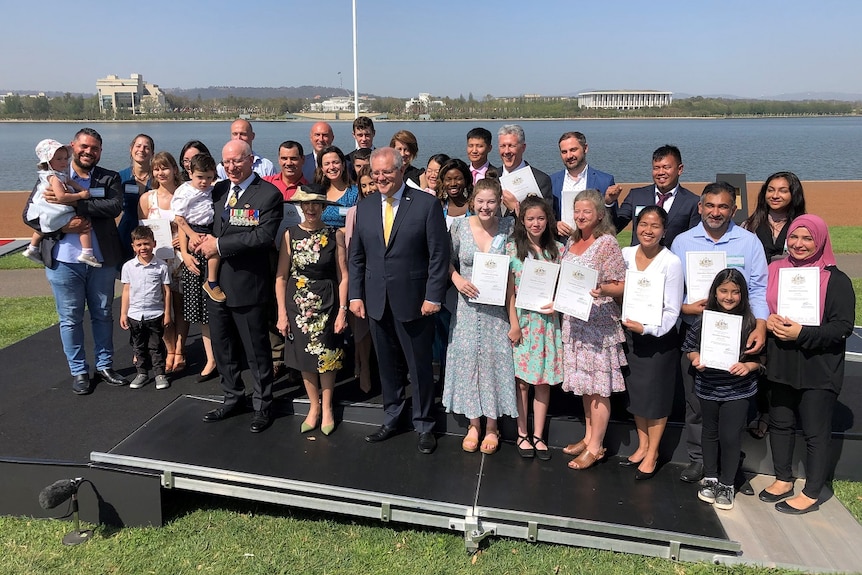 A group of people who have just attended their citizenship ceremony stand with Prime Minister Scott Morrison next to a lake