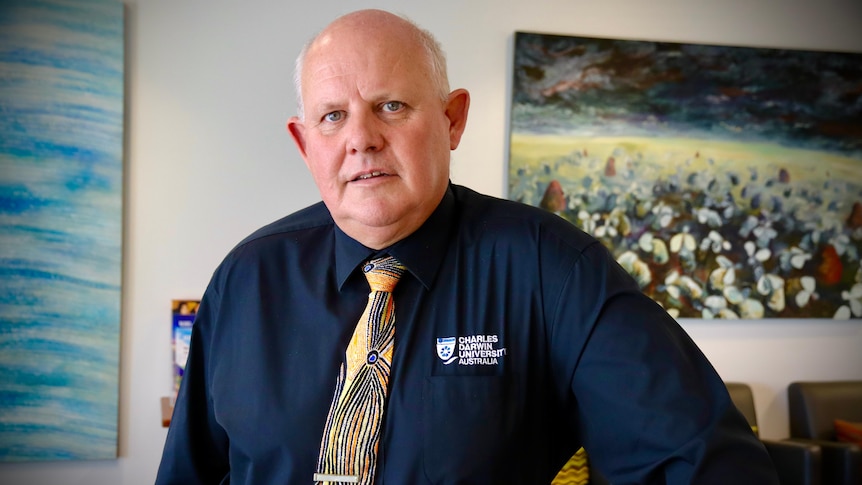An older balding white man wearing a Dark blue CDU shirt and tie in an Aboriginal pattern stands in his office.