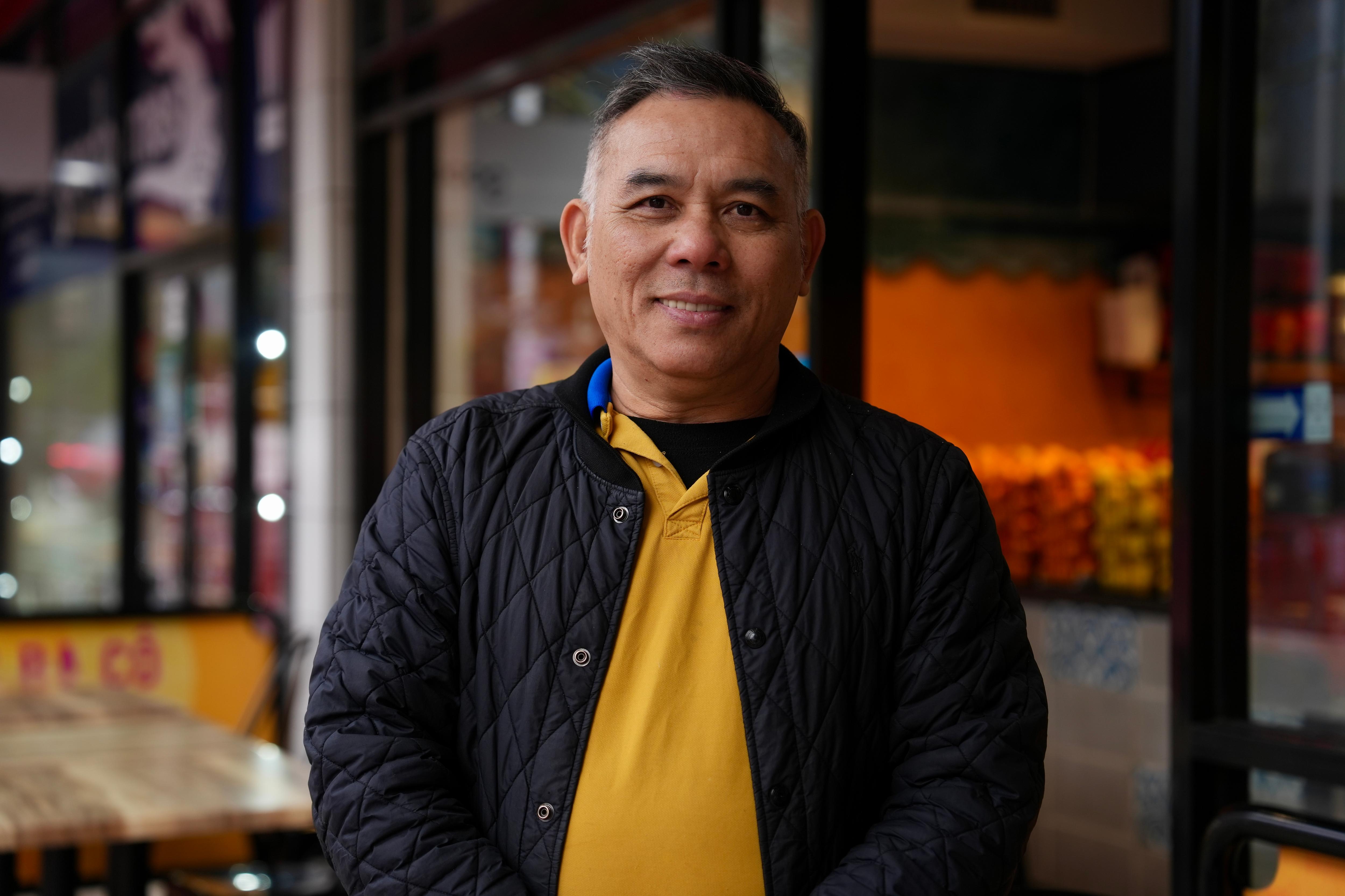 Richard sonríe, parado fuera de su negocio con una chaqueta y un jersey.