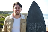 A man with short brown hair wearing a tan-coloured corduroy jacked stands at a beach holding a black surfboard.