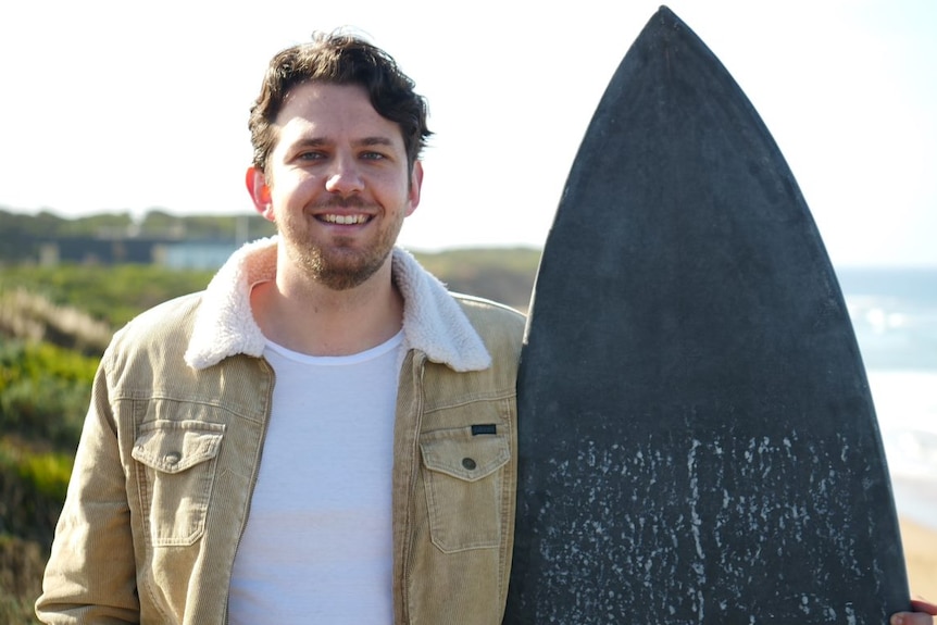 A man with short brown hair wearing a tan-coloured corduroy jacked stands at a beach holding a black surfboard.