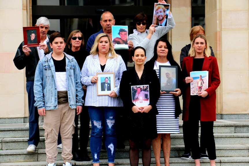 A group of family members of people killed in the workplace stand on the steps of the WA Parliament holding framed photographs.