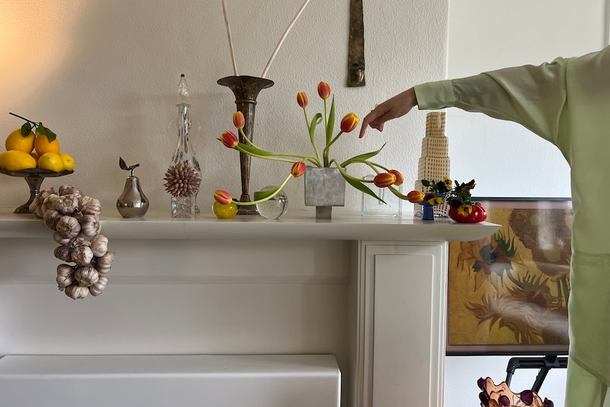 A hand points at a silver square vase on a mantelpiece with red-orange tulips in it, surrounded by trinkets.