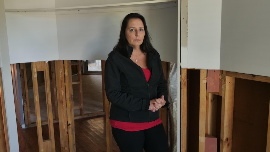 A woman wearing a jacket looks seriously at the camera inside her damaged home.