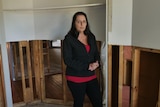 A woman wearing a jacket looks seriously at the camera inside her damaged home.